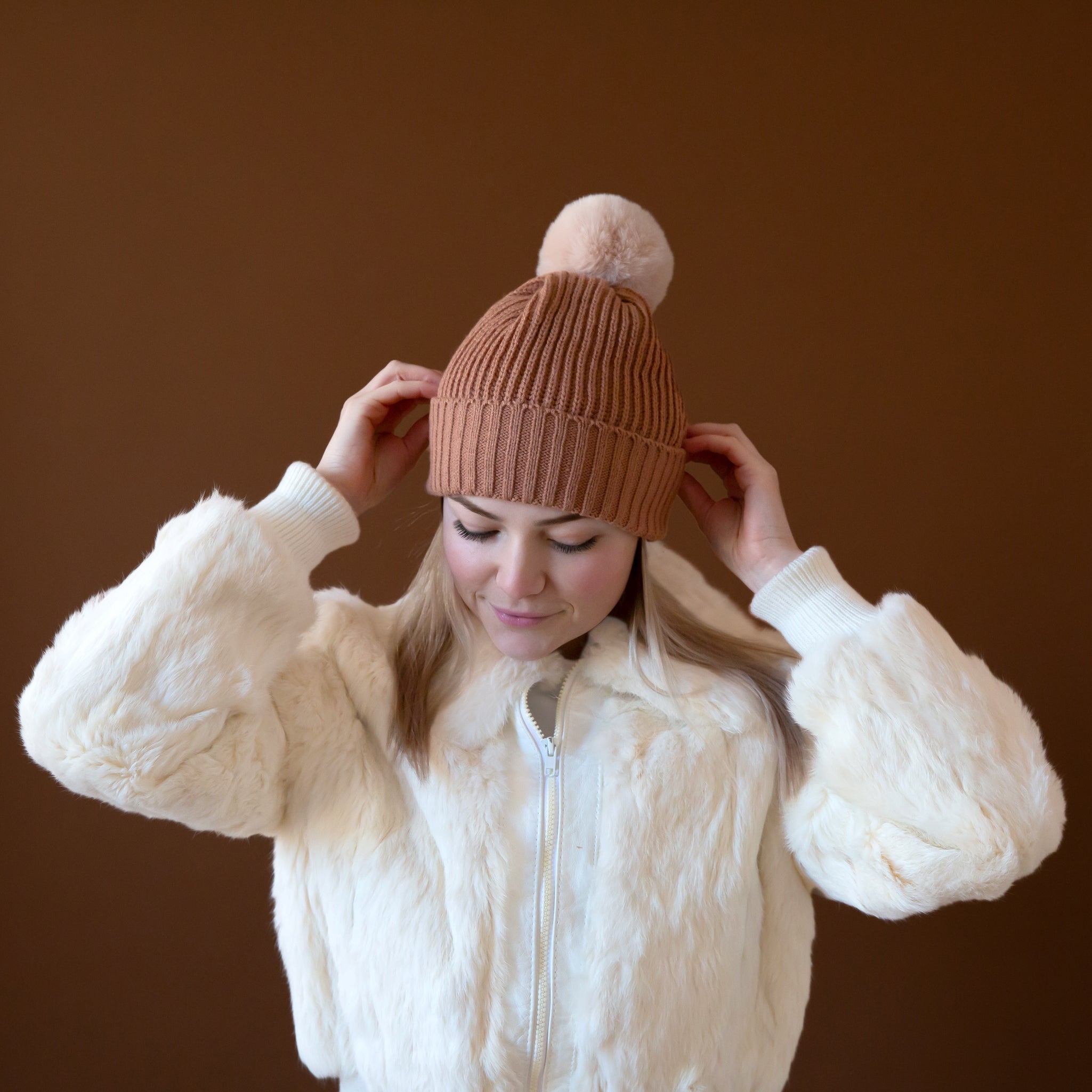 A model wearing a light brown beanie with a neutral pom at the top. 