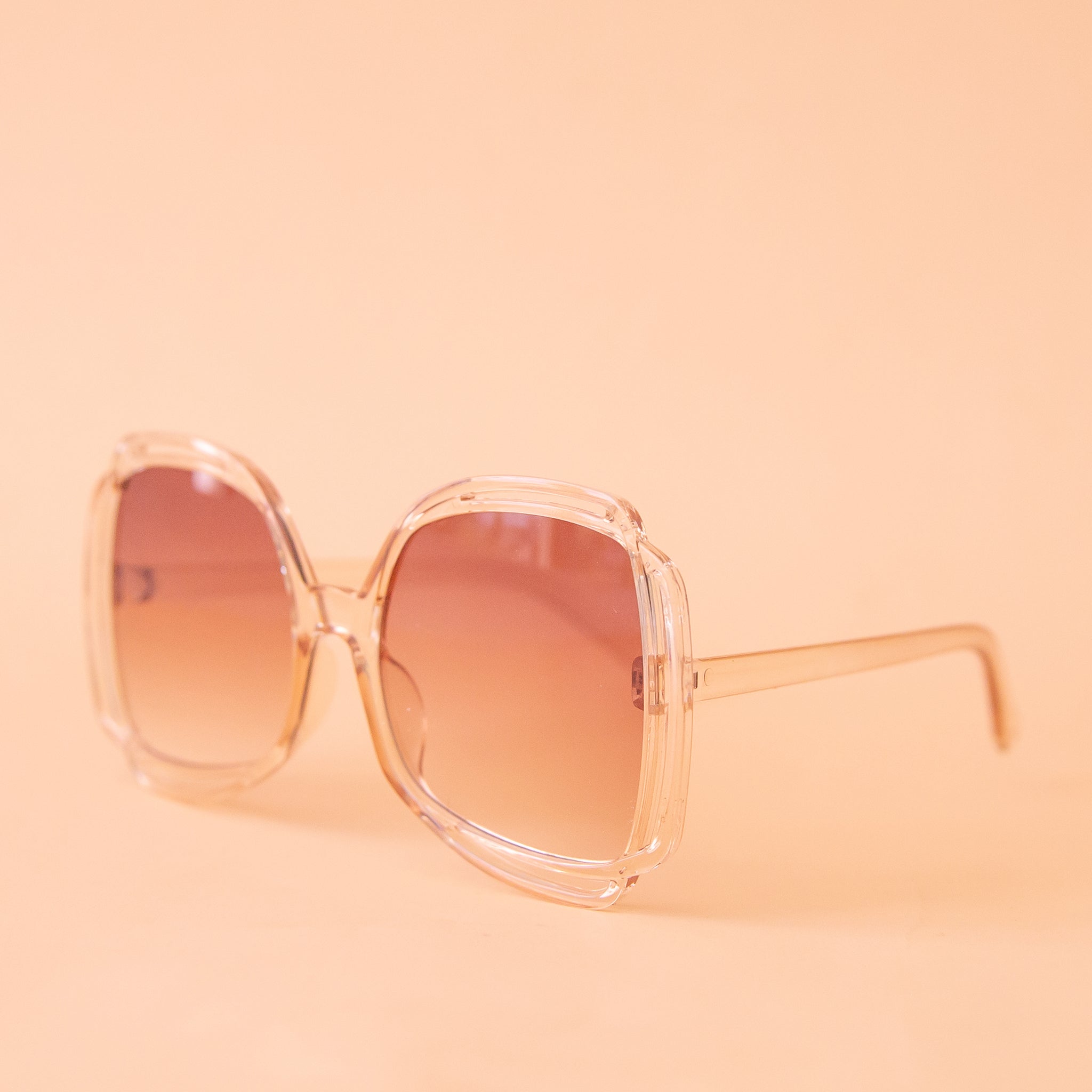 A pair of sunglasses with a clear peachy frame and pinkish gradient lenses.