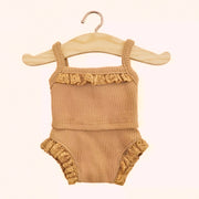 On a light tan background is a ribbed two piece doll outfit that features a ruffle detail around the legs and the neckline. The outfit is a soft brown color and hangs on a wooden hanger.