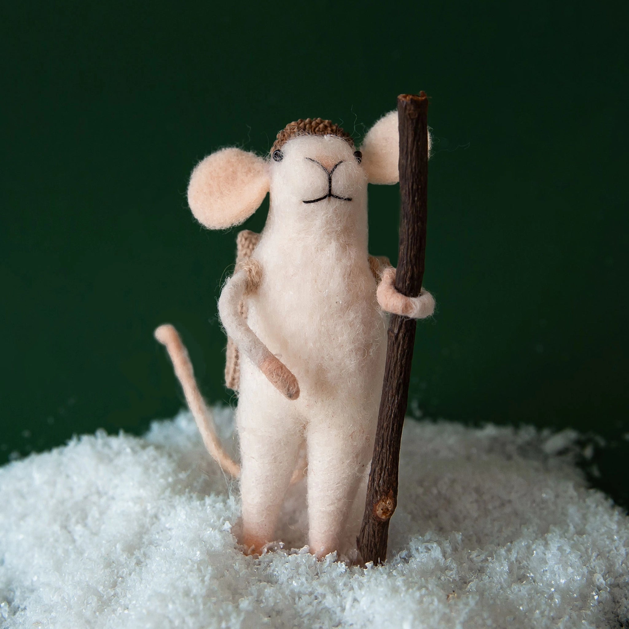 On a green background is a white felt mouse ornament with a wood walking stick and an acorn hat. 