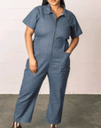 On a neutral background is a model wearing a denim short sleeved jumpsuit with a front zipper and a collar. 