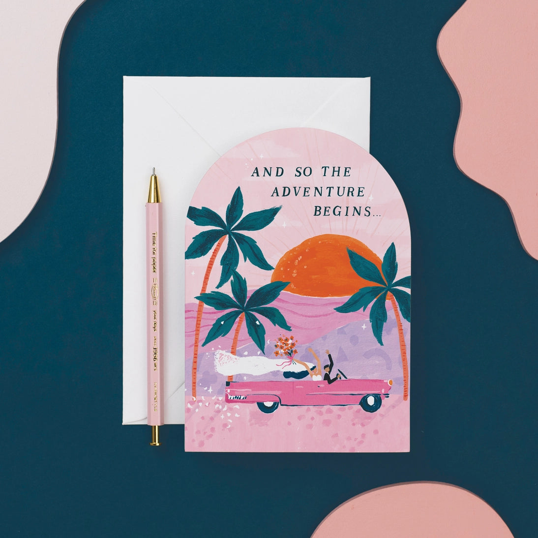 A pink arched greeting card featuring three palm tree illustrations along with a pink vintage car riding into the sunset with a bride and groom holding their hands and bouquets in the air as well as text that reads, &quot;And So The Adventure Begins...&quot; in black letters.