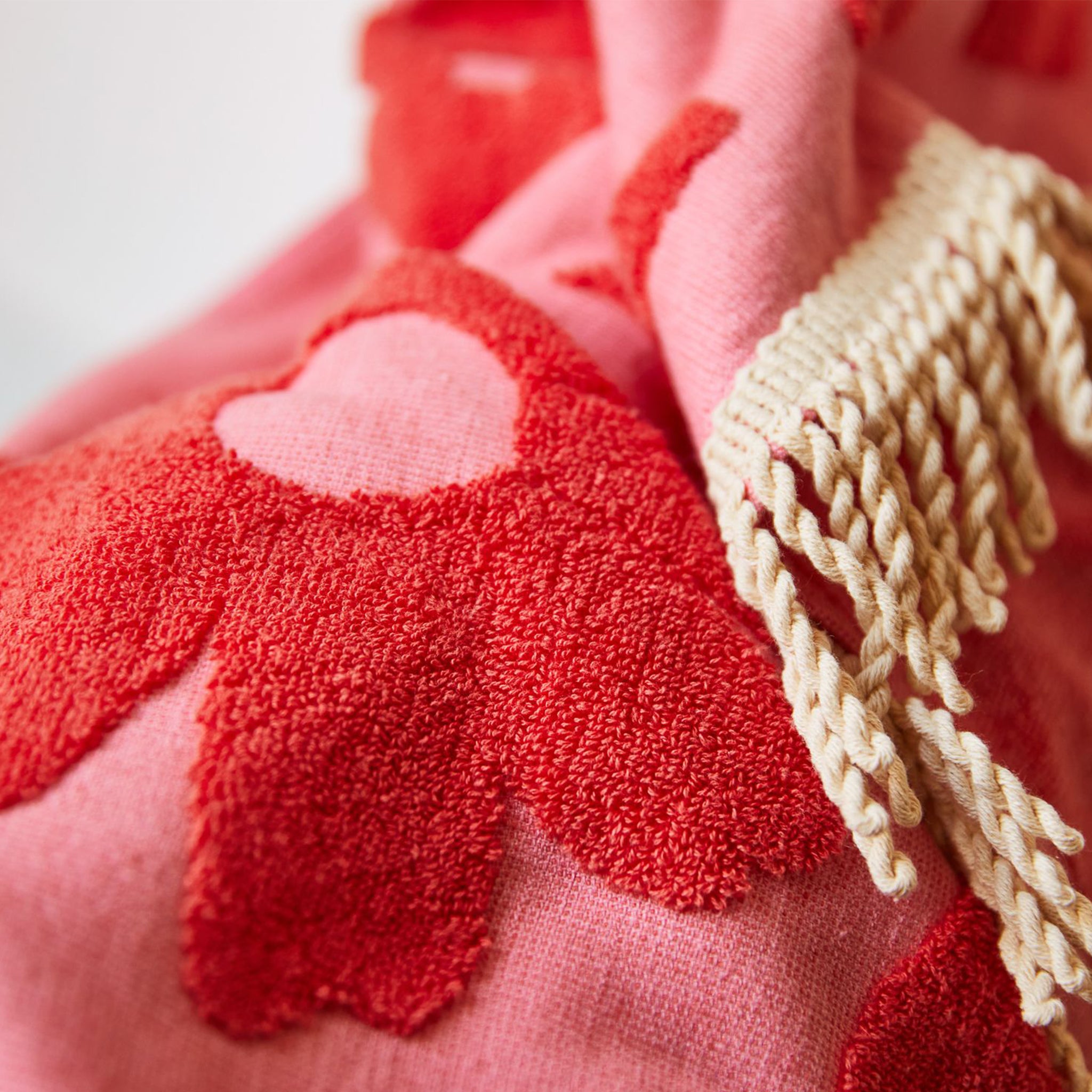 A light and dark pink hand towel with a floral pattern and fringe detailing on two ends. 