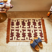 A cream, brown and red geometric patterned bath mat with tassel edges on two sides. 