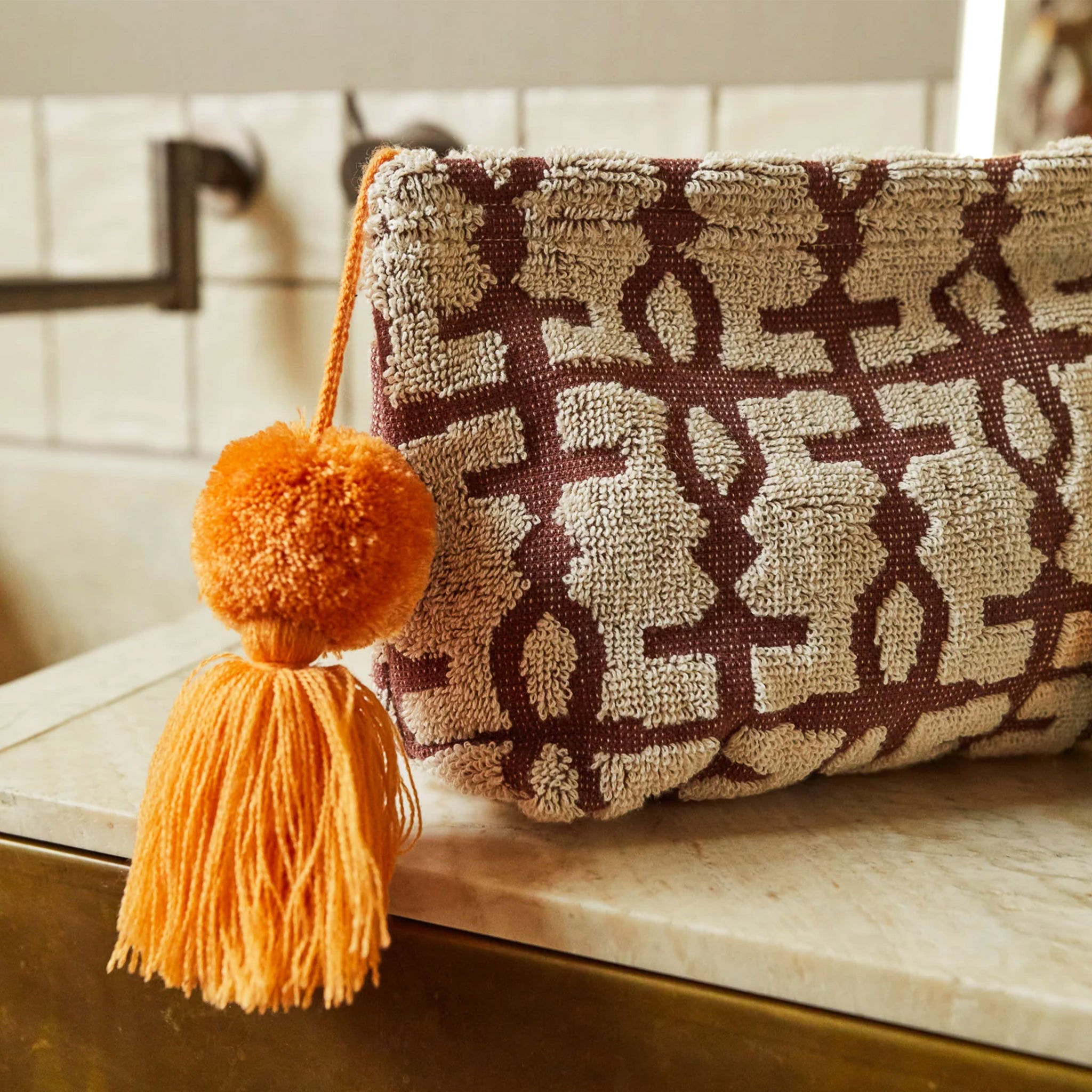 A tan and brown geometric designed terry cosmetic pouch in two different sizes with an orange tassel on the zipper. 