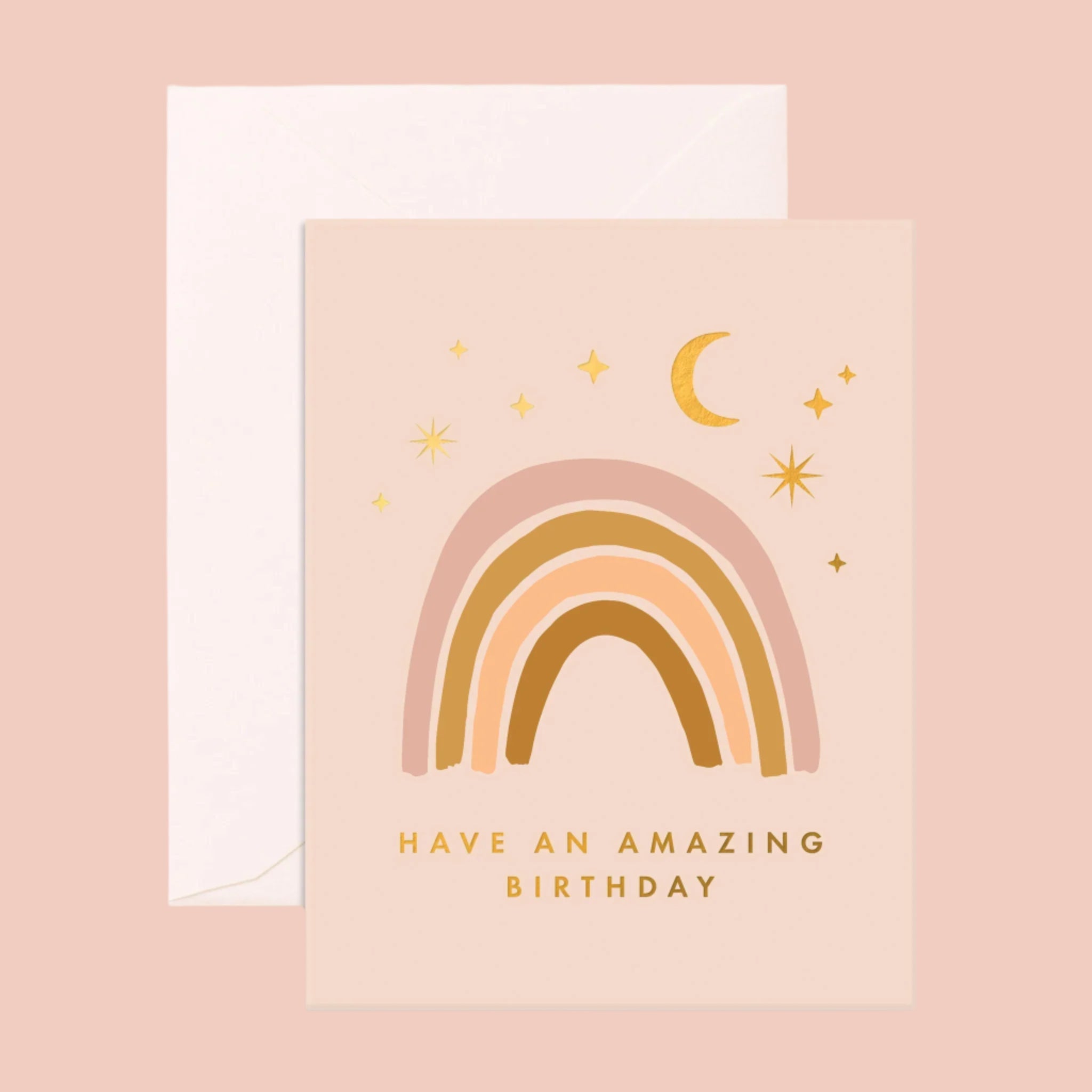 A light pink card with a neutral rainbow on the front under moon and star designs and gold text along the bottom that reads, &quot;Have an amazing birthday&quot; as well as a coordinating white envelope.