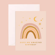 A light pink card with a neutral rainbow on the front under moon and star designs and gold text along the bottom that reads, "Have an amazing birthday" as well as a coordinating white envelope.