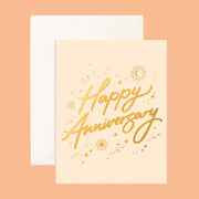 On a neutral background is a tan card with gold foil text that reads, "Happy Anniversary" along with gold foil stars, sun and moons around. 