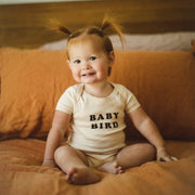 Toddler with pigtails sitting and smiling on an orange couch, wearing a "BABY BIRD" onesie.