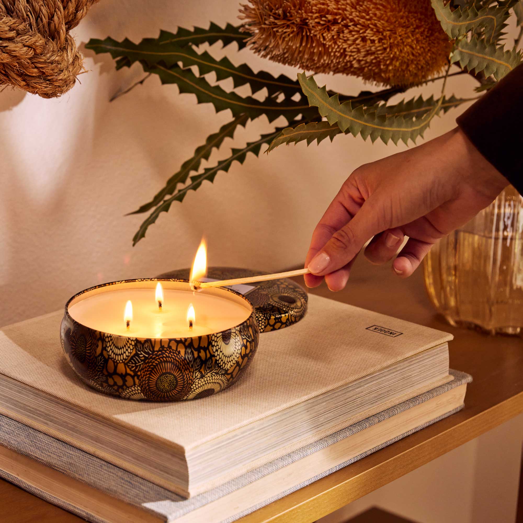 Round tin candle vessel with dark and light gold floral pattern and white wax candle with three wicks. Candle vessel lid has text, "Voluspa. Baltic Amber."