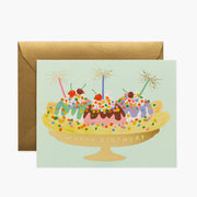 On a white background is a mint green birthday card with an illustration of a banana split along with gold foiled text that reads, "Happy Birthday".