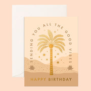 Warm toned birthday card complete with a desert scene. The text 'Sending you all the good vibes' curves around a gold foil palm tree and desert stars. Below the scene reads 'Happy Birthday' in capital gold foil lettering. The card is accompanied by a solid white envelope.