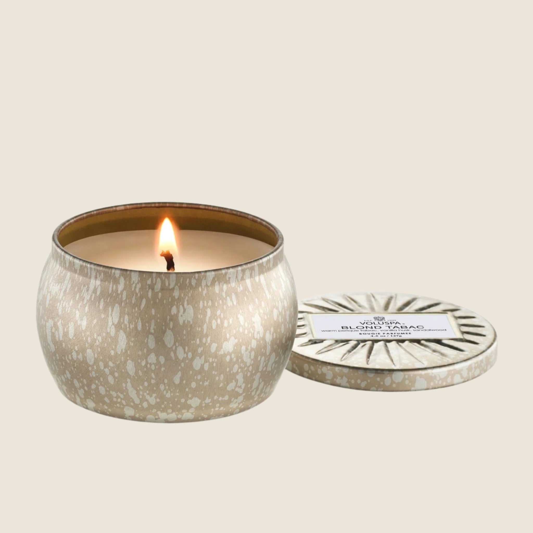 On a cream background is a light silver and gold mini tin candle with white wax and a single wick. Also included is the matching lid.