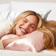 A smiling woman with long blonde hair rests her head on a blush colored satin pillow.