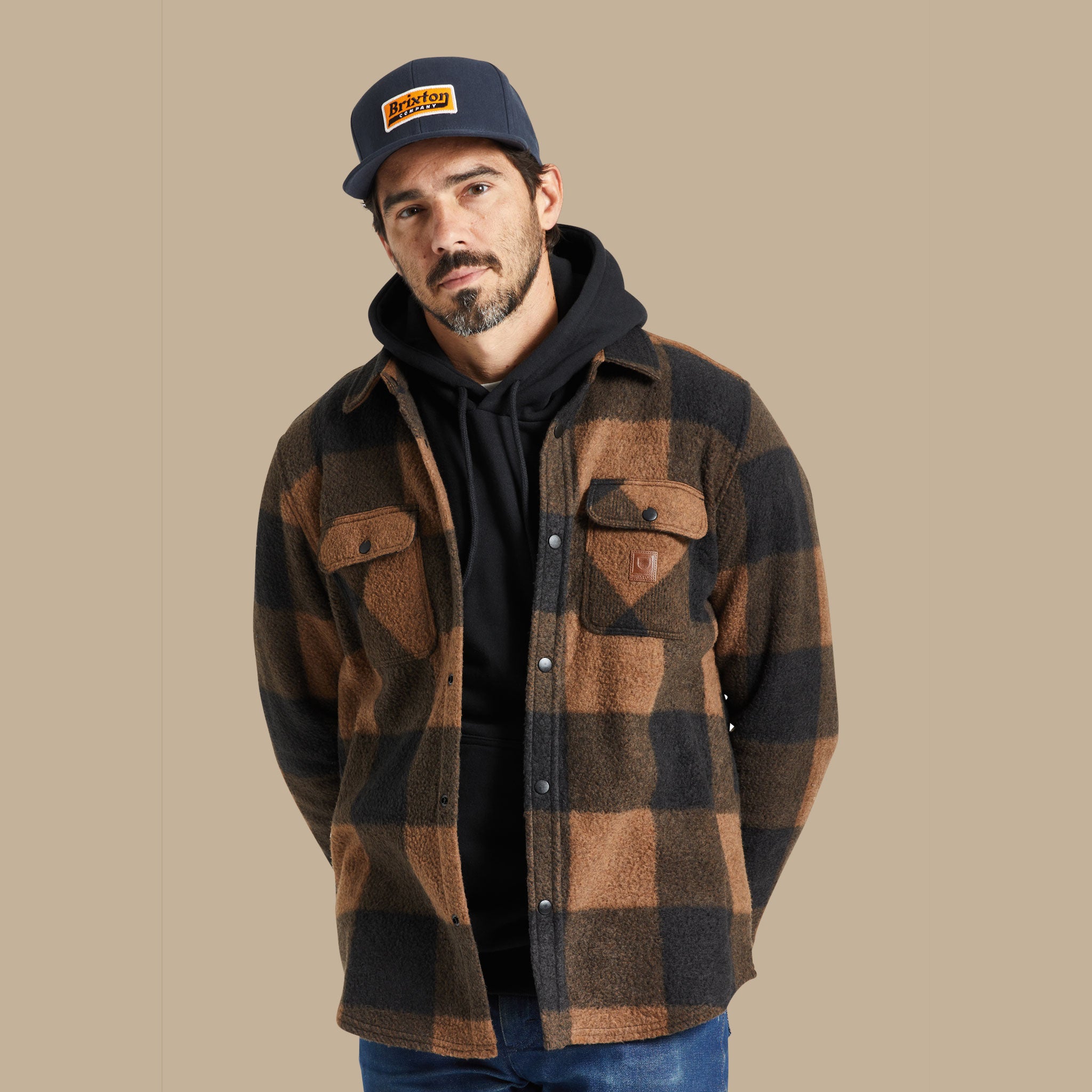 On a tan background is a male model wearing a black and brown plaid fleece shacket with front pockets and buttons. 