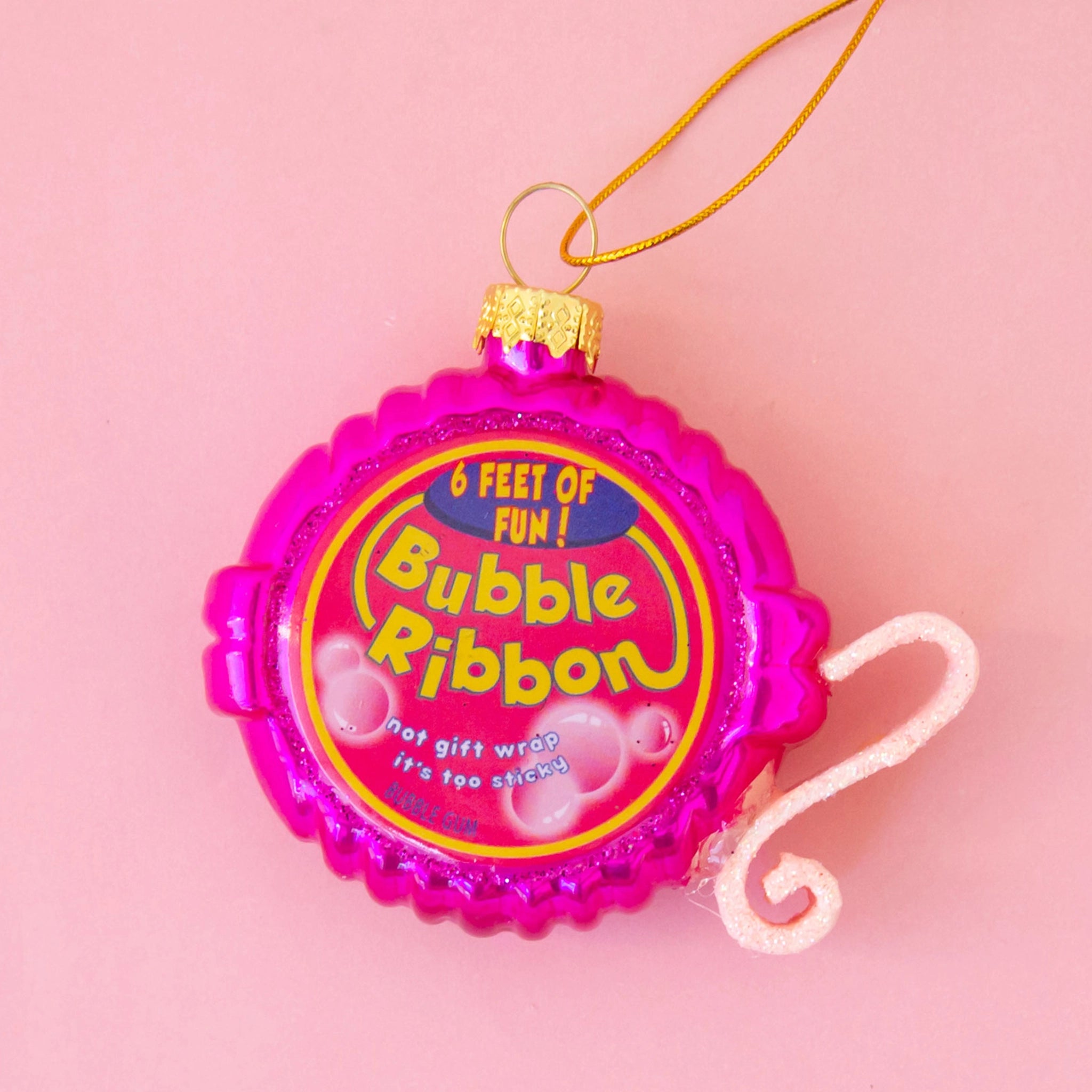 On a pink background is a bright pink Bubble Gum Tape Dispenser ornament with a gold loop for hanging.