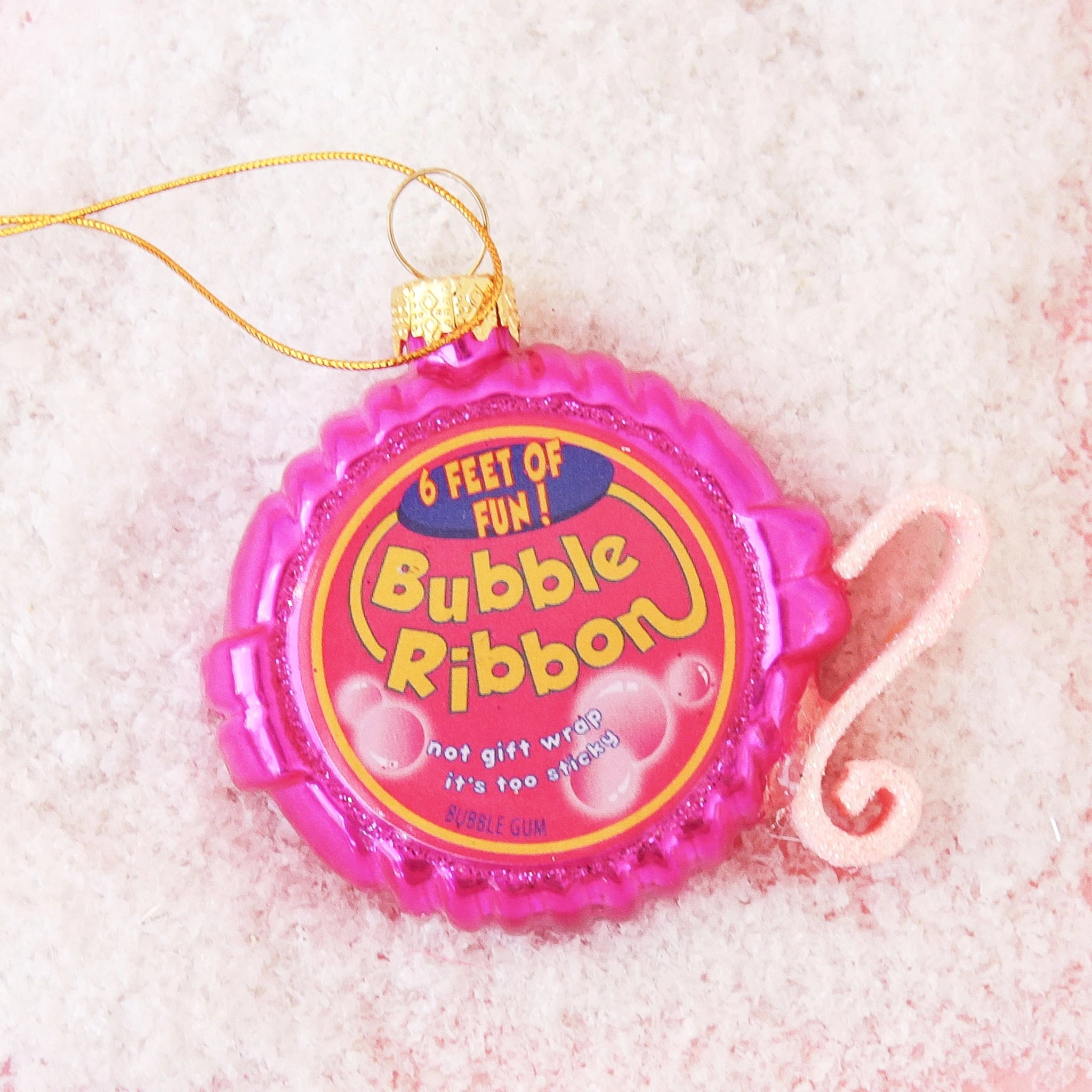 On a pink background is faux snow, is a bright pink Bubble Gum Tape Dispenser ornament with a gold loop for hanging.  