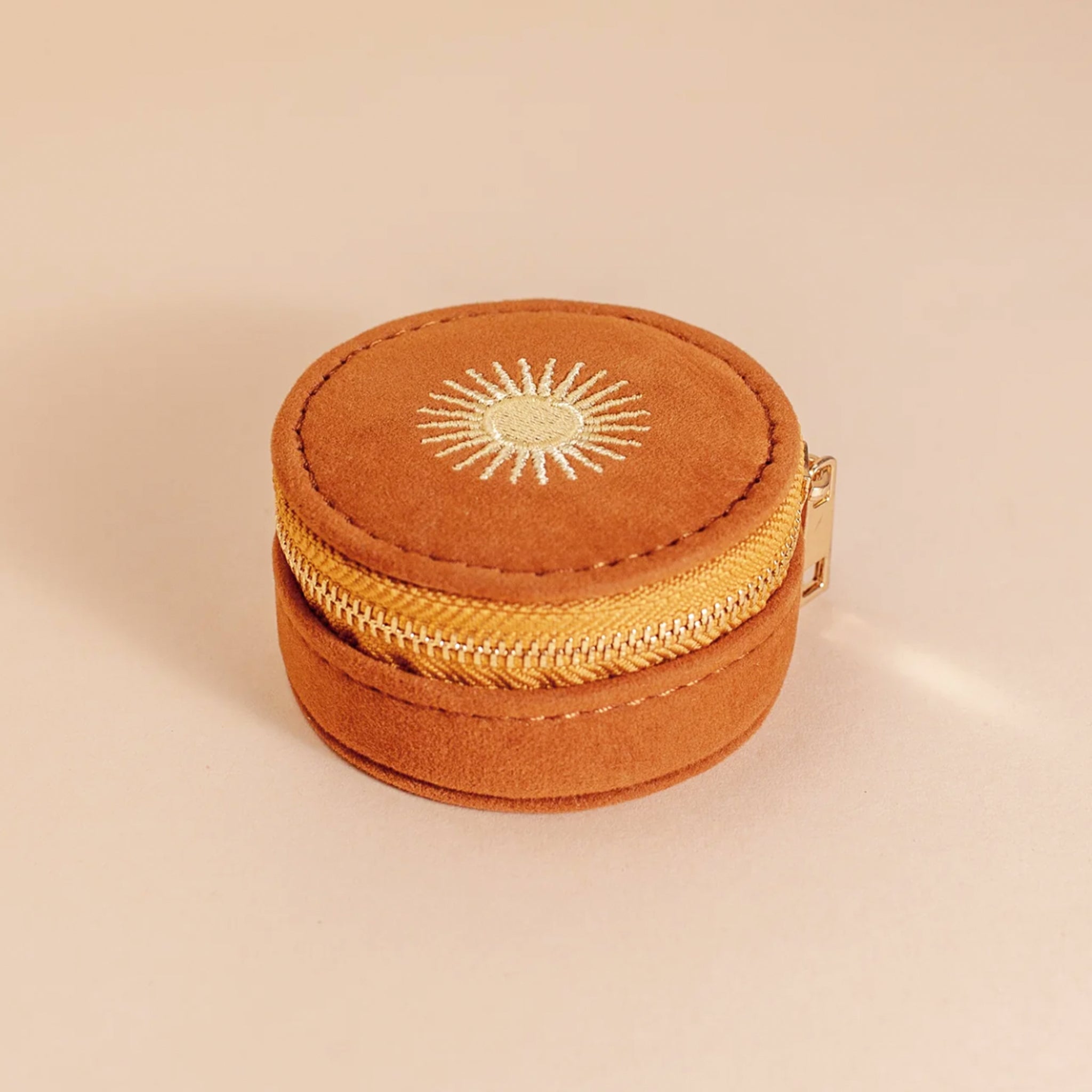 A circular suede burnt orange jewelry case with a gold zipper and a sun design on top. 