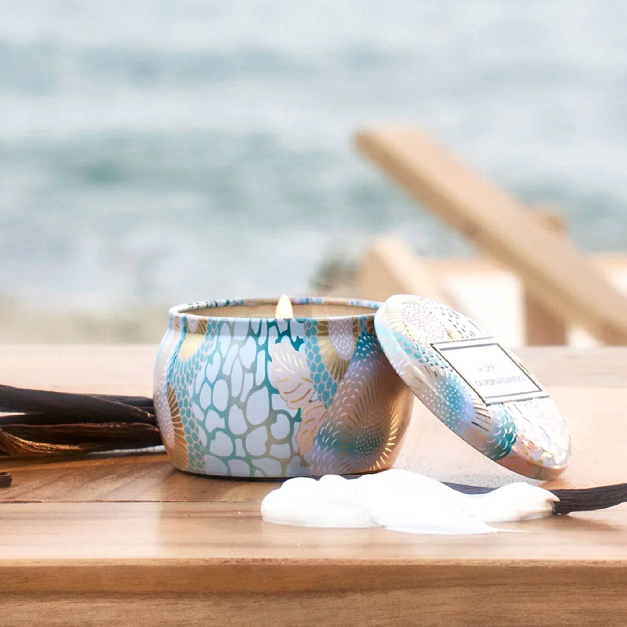 On a blue background is a light blue decorative mini tin candle with a lid and a rectangle label that reads, "Voluspa California Summers". 