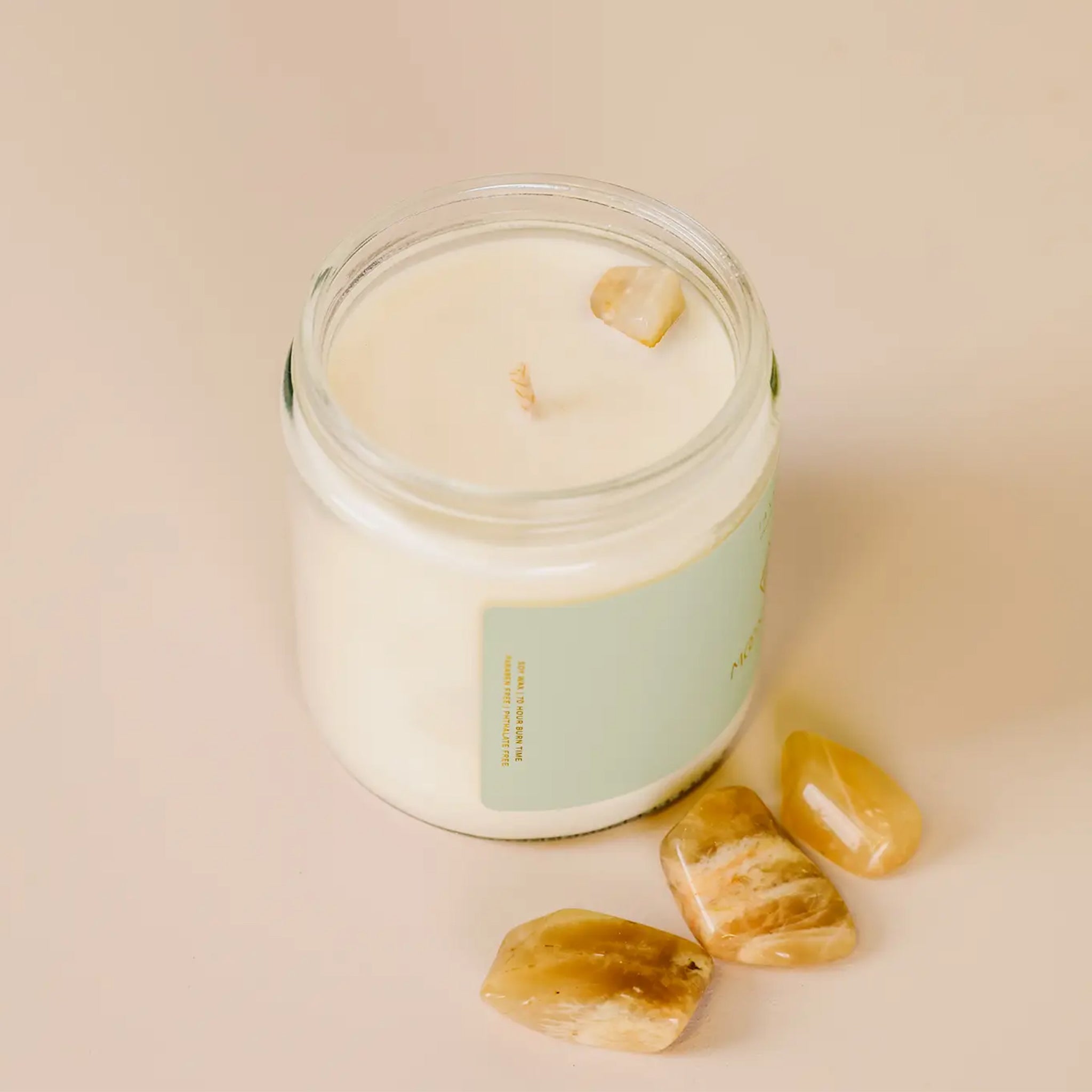 On a tan background is a clear glass jar candle with a light blue label and gold lid.