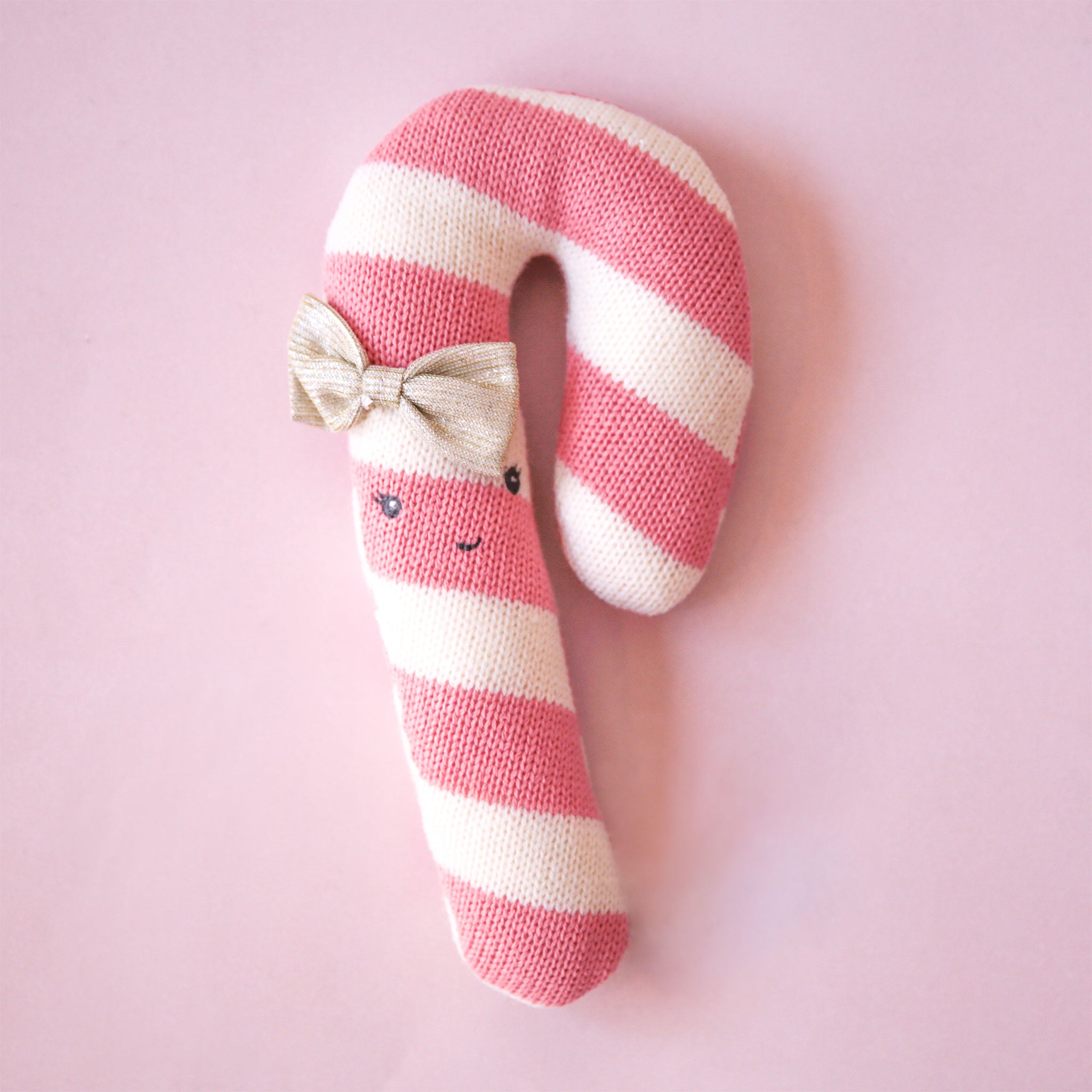 On a pink background is a pink and white candy cane stuffed toy with an embroidered smile and neutral bow. 