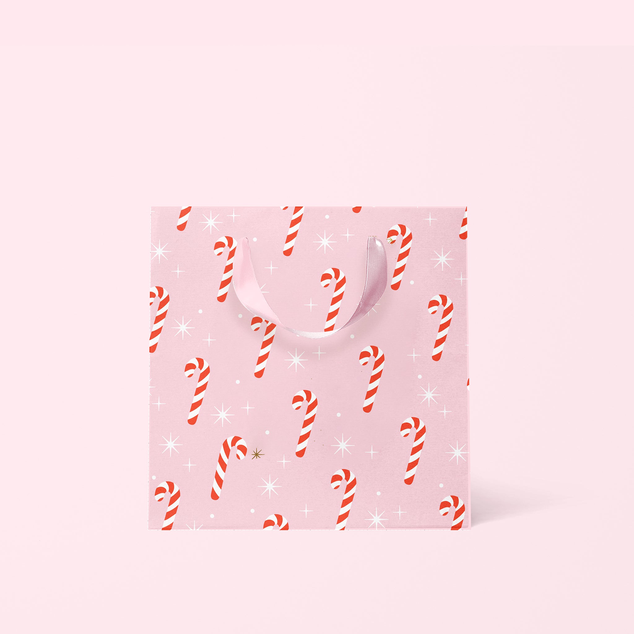 Digital mockup of the small candy cane gift bag. The gift bag features red and white candy canes on a pink ground surrounded by white sparkles. The bag sits on a pink paper ground. 