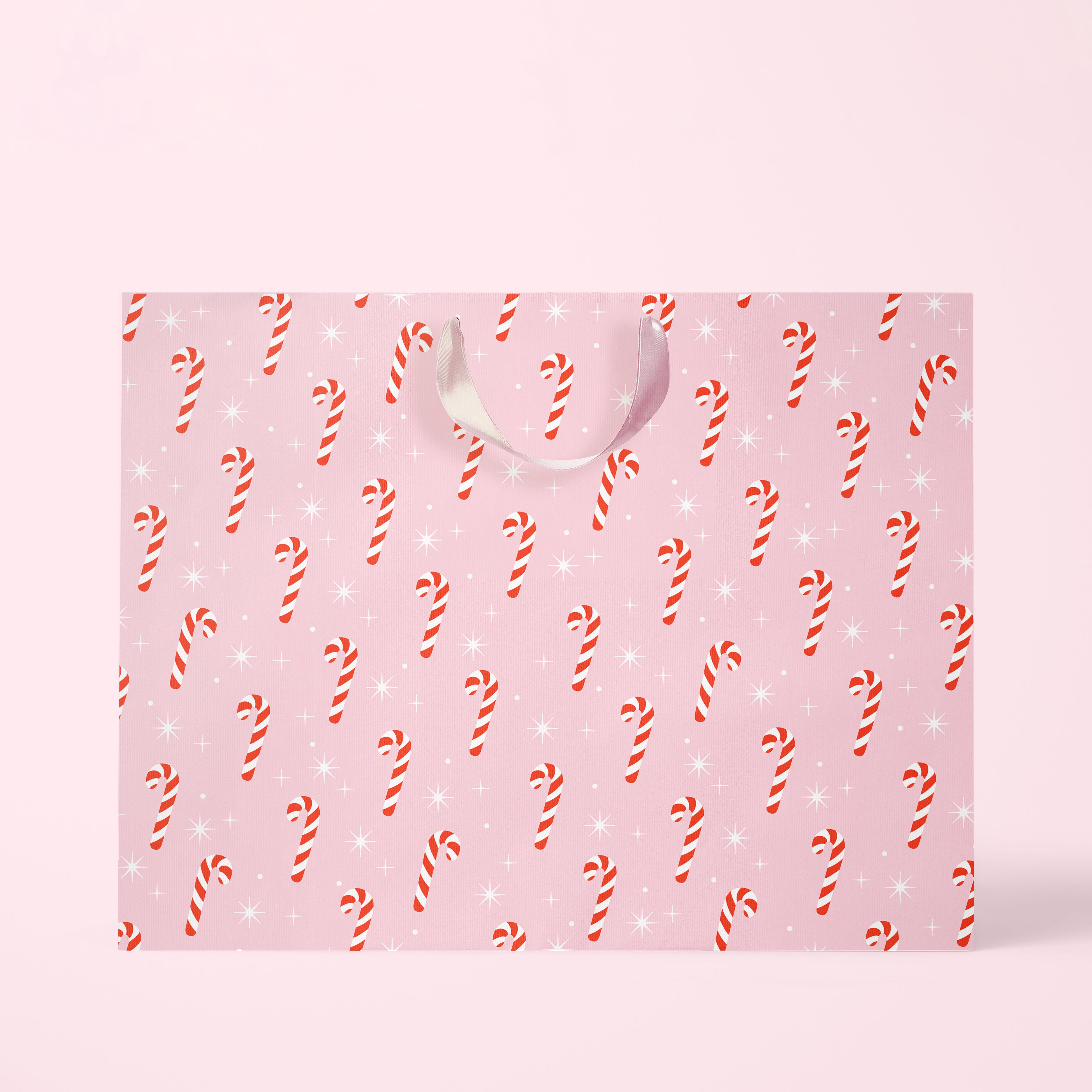 Digital mockup of the large candy cane gift bag. The gift bag features red and white candy canes on a pink ground surrounded by white sparkles. The bag sits on a pink paper ground. 