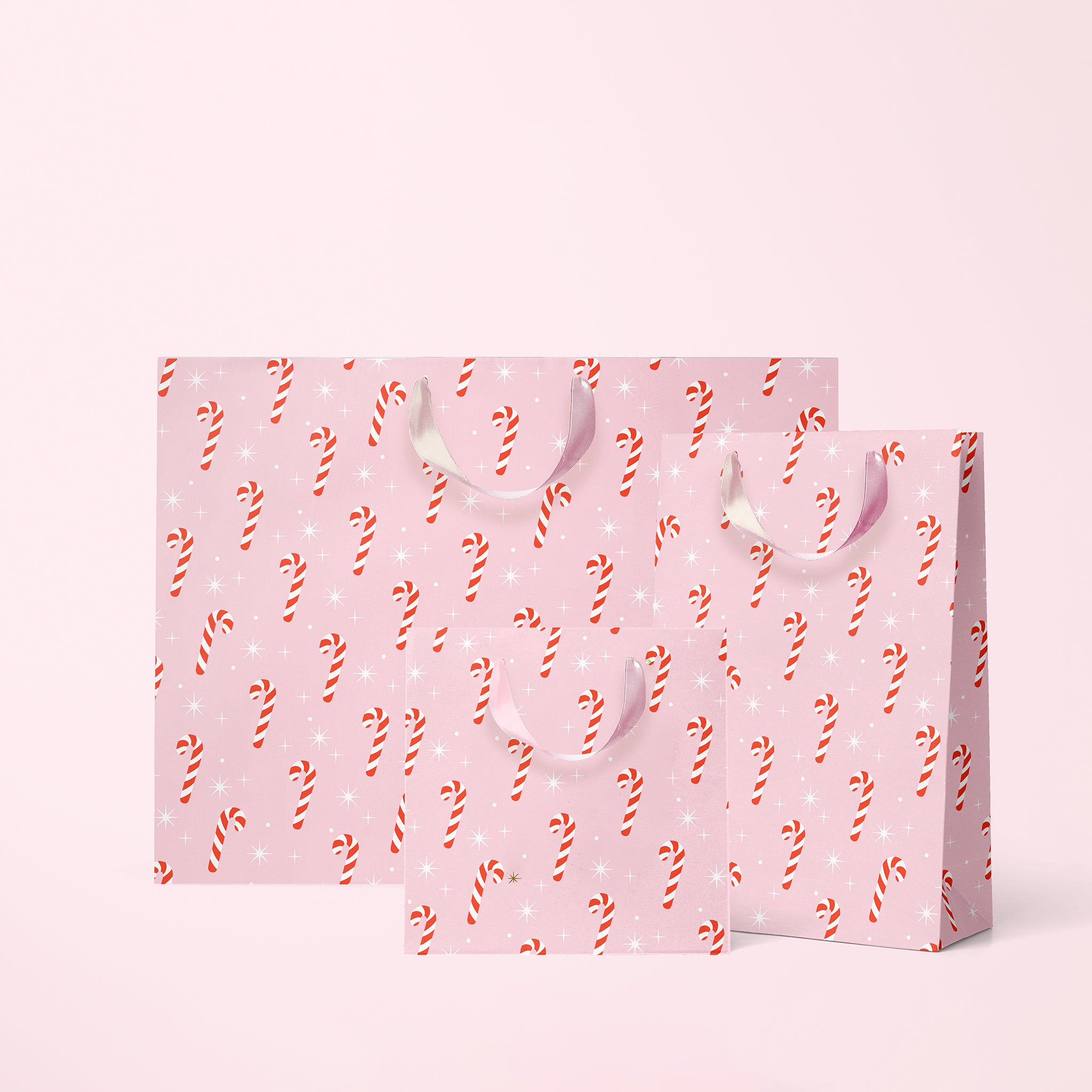 Digital mockup of three gift bags, small, medium and large that feature a pink background with red and white candy canes surrounded by white sparkles. they have a ribbon handle and sit on a pink paper background. 