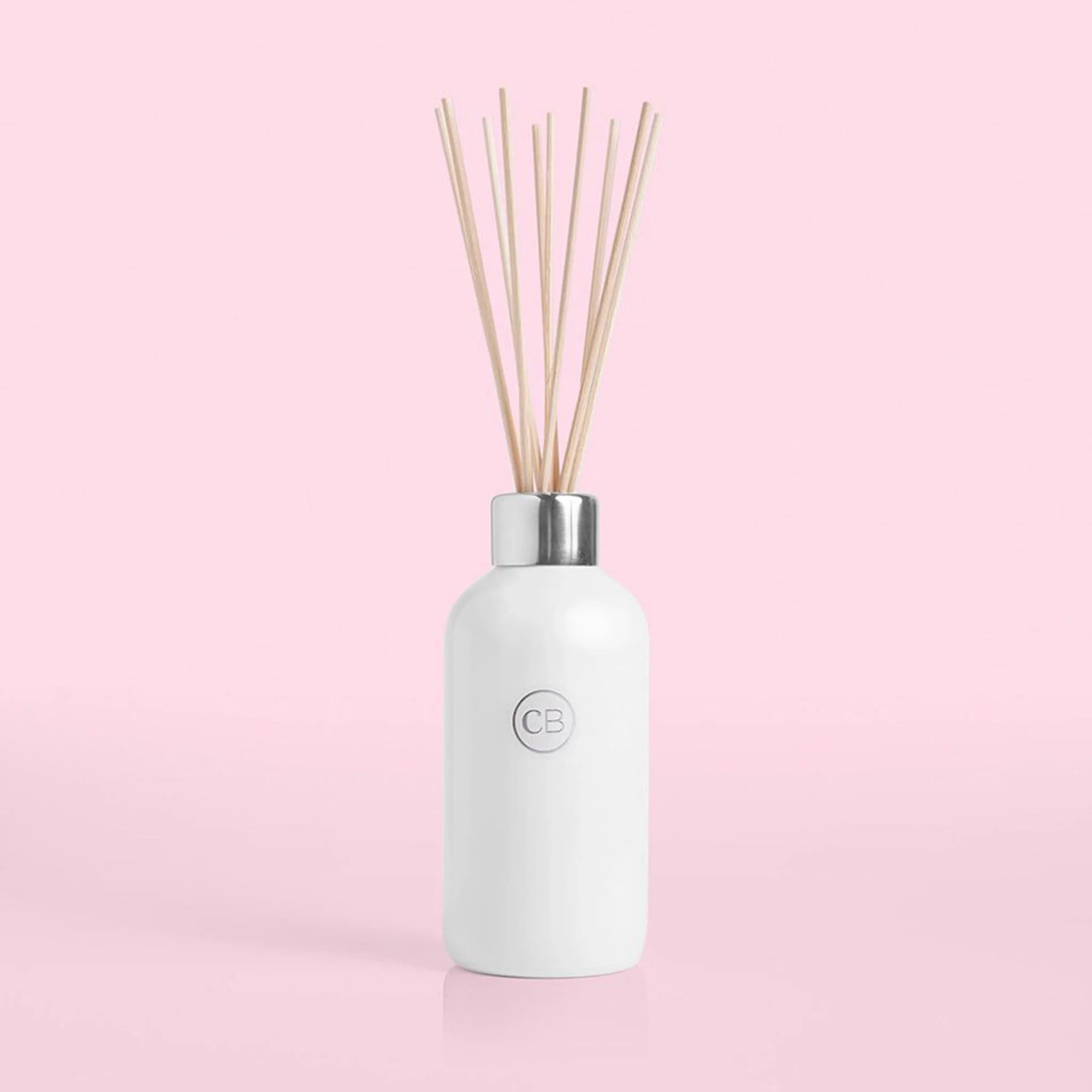 On a pink background is a white glass diffuser jar with a silver lid and multiple wooden reeds coming out of the base that holds the scented liquid.