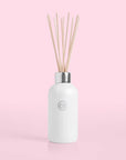On a pink background is a white glass diffuser jar with a silver lid and multiple wooden reeds coming out of the base that holds the scented liquid.
