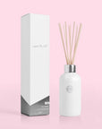 On a pink background is a white glass diffuser jar with a silver lid and multiple wooden reeds coming out of the base that holds the scented liquid. Also photographed here is the white and silver box that the diffuser comes in that reads, "Capri Blue" in silver letters.