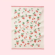 A tea towel with red cherries all over with green leaves lays in the middle of page.  there is a soft pink background and the towel has bright red ric-rac lining the sides.