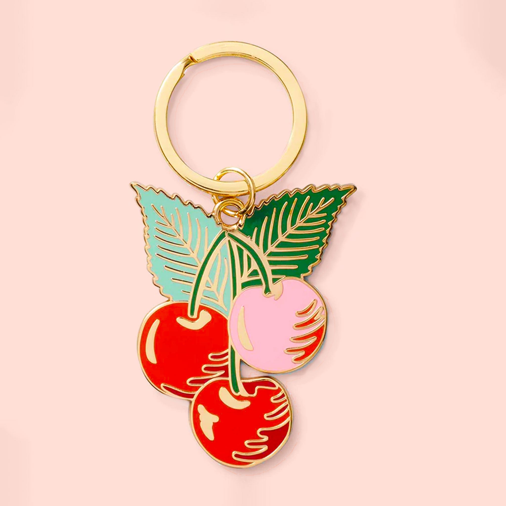 A gold keychain with three cherries and two leaves sits on a pink background.  The cherries are red and pink and the leaves are mint and spearmint green.