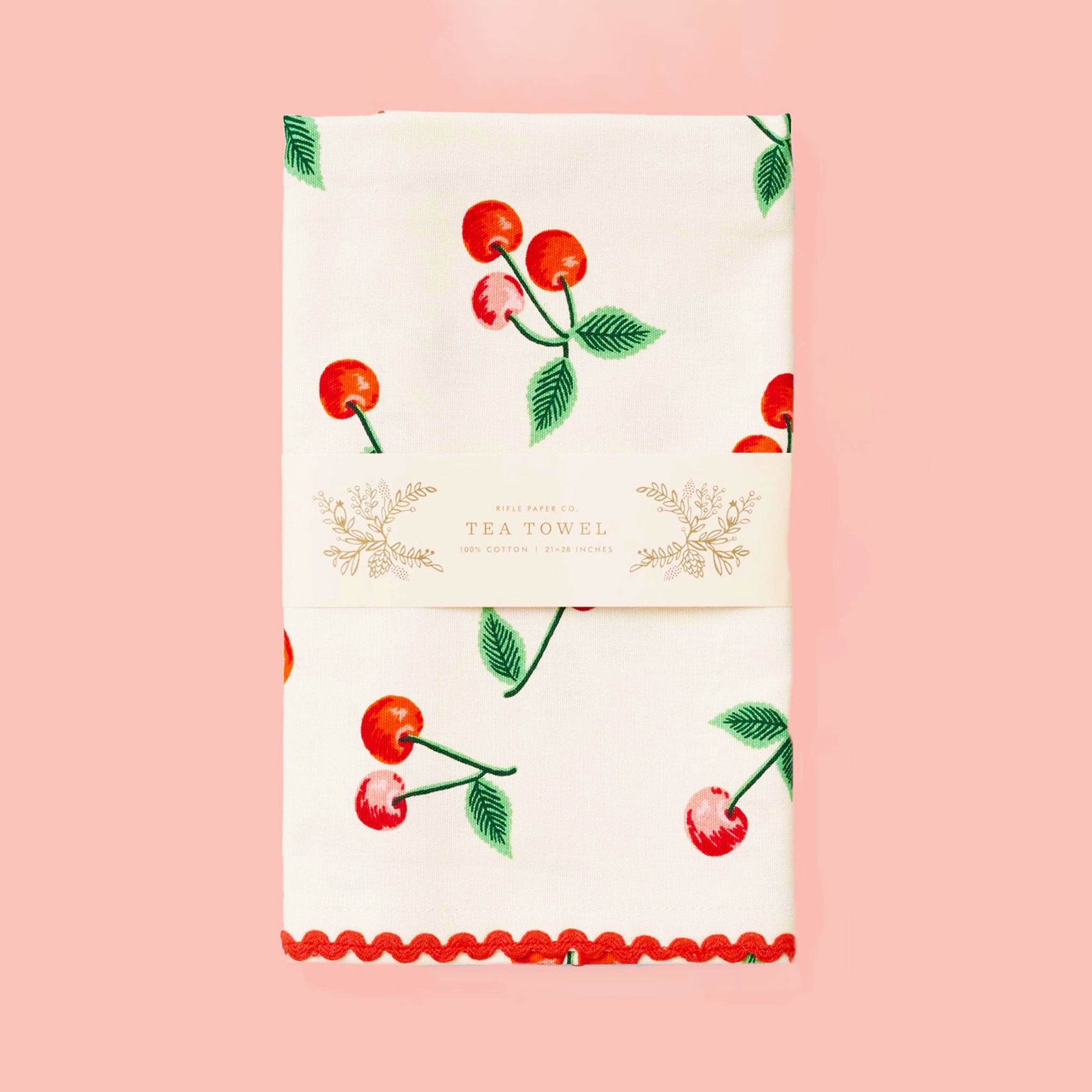 a folded tea towel sits on a pink background.  The towel is a white color with red cherries with green leaves scattered across the fabric.  There is a band of paper across the middle of the folded towel that reads &quot;tea towel&quot;.