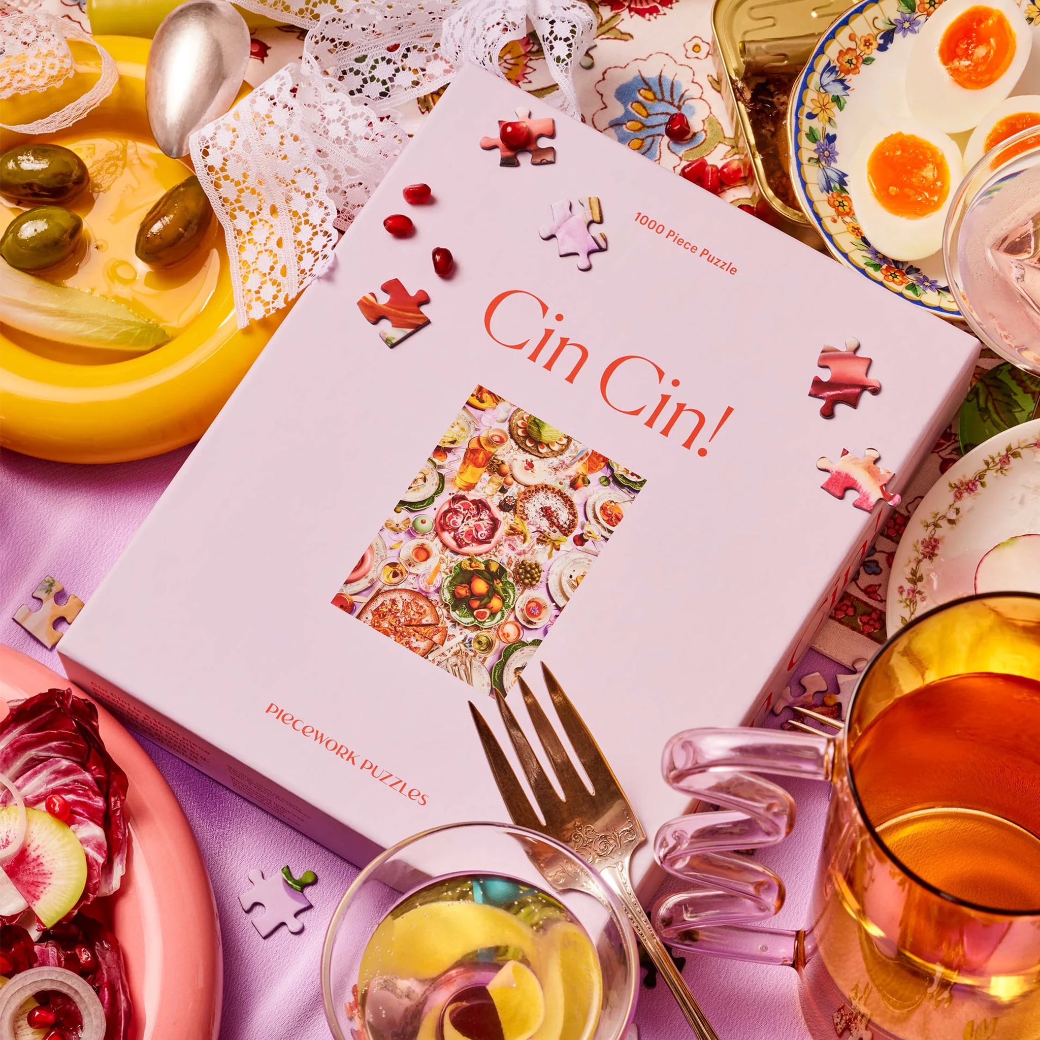 The back of the box that read features a colorful tablescape of drinks and food along with red text along the side that reads, "Piecework Puzzles". 