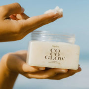 A clear container of a white body scrub with a white lid and black text on the front that reads, "CocoGlow". 