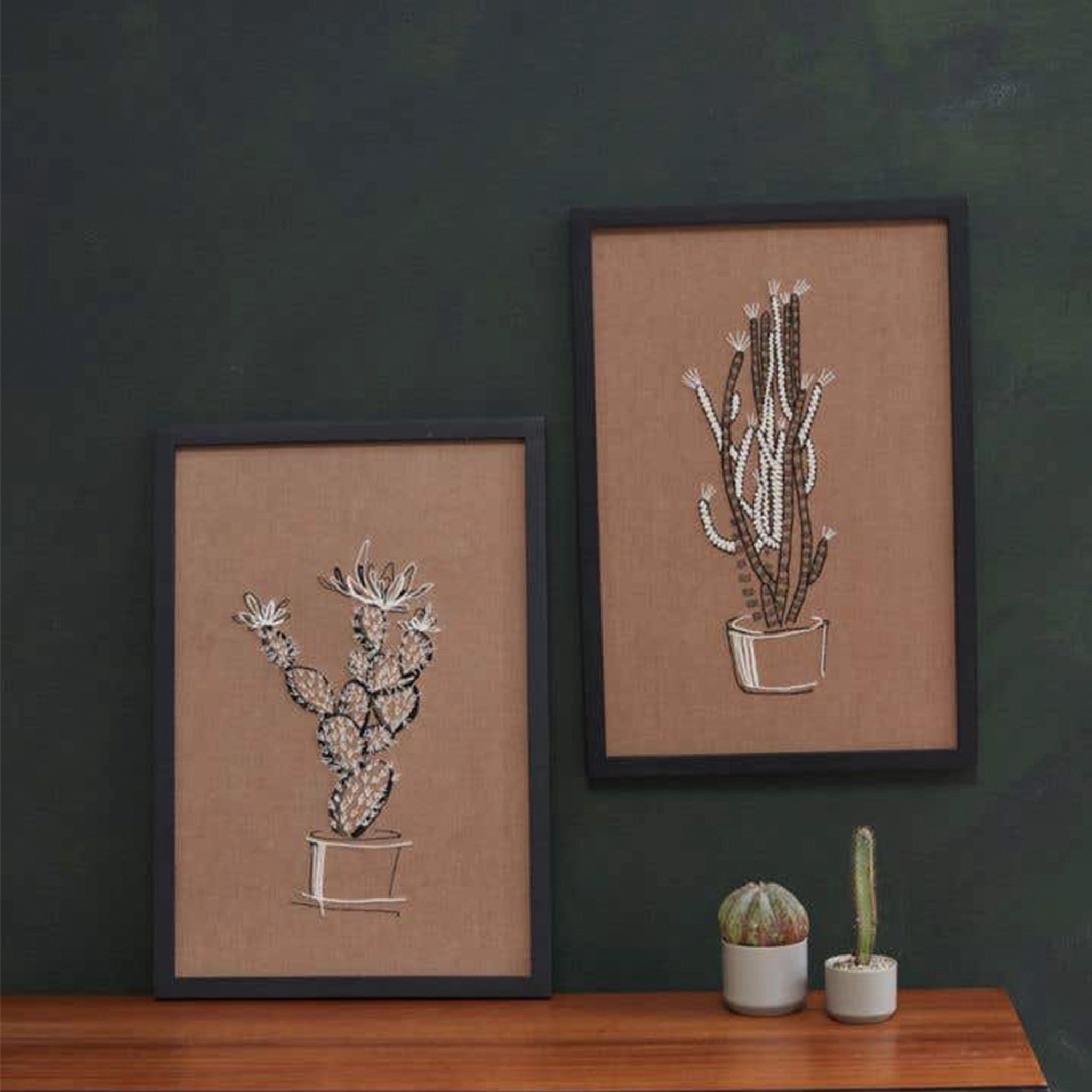 On a white background is a brown piece of wall art with white and black stitching of a cactus in a pot. The art comes in a simple black frame. In this photo, the Prickly Pear wall art is staged next to another piece of wall art. 