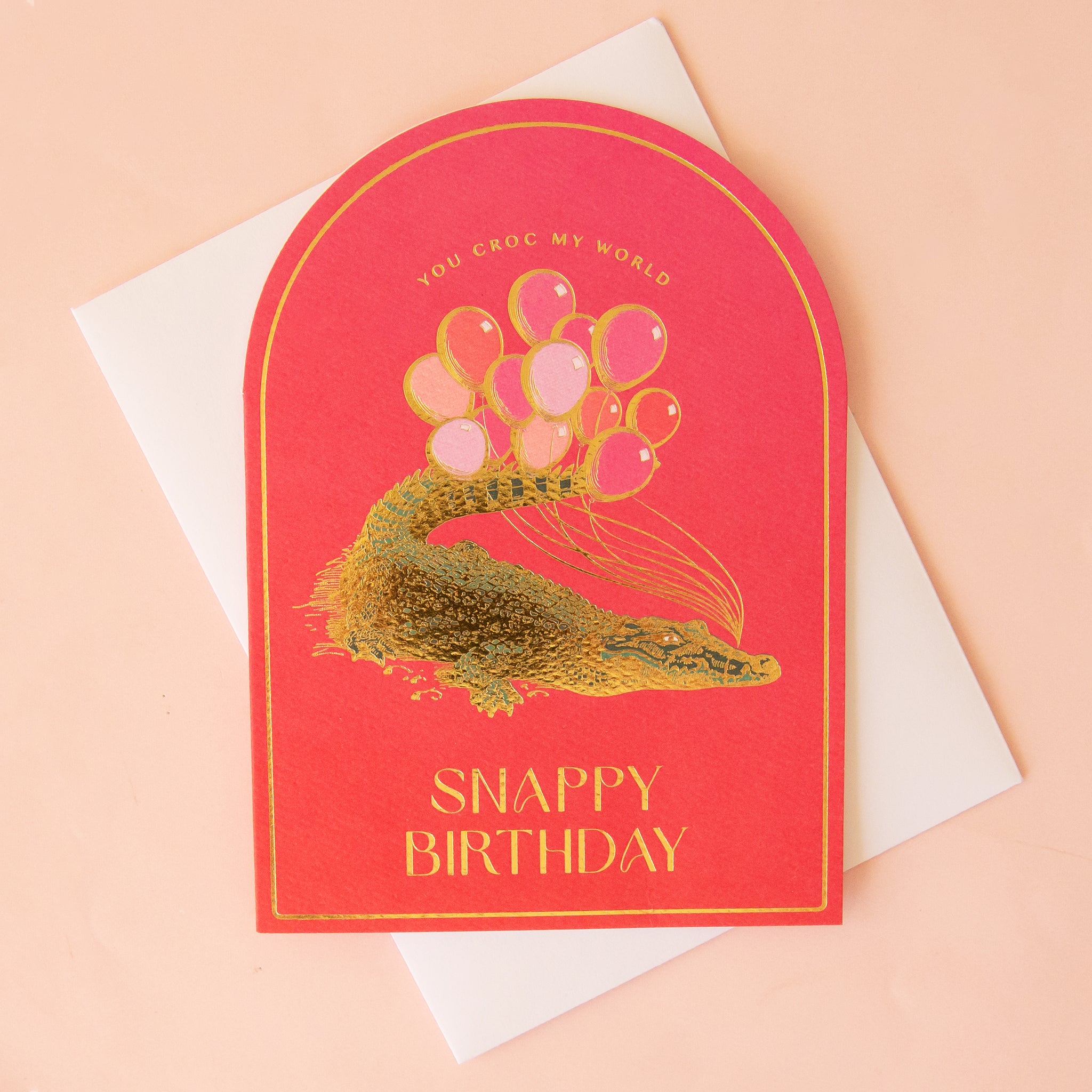 A hot pink arched card with a graphic of a crocodile holding balloons and text that reads, &#39;You Croc My World Snappy Birthday&quot;.