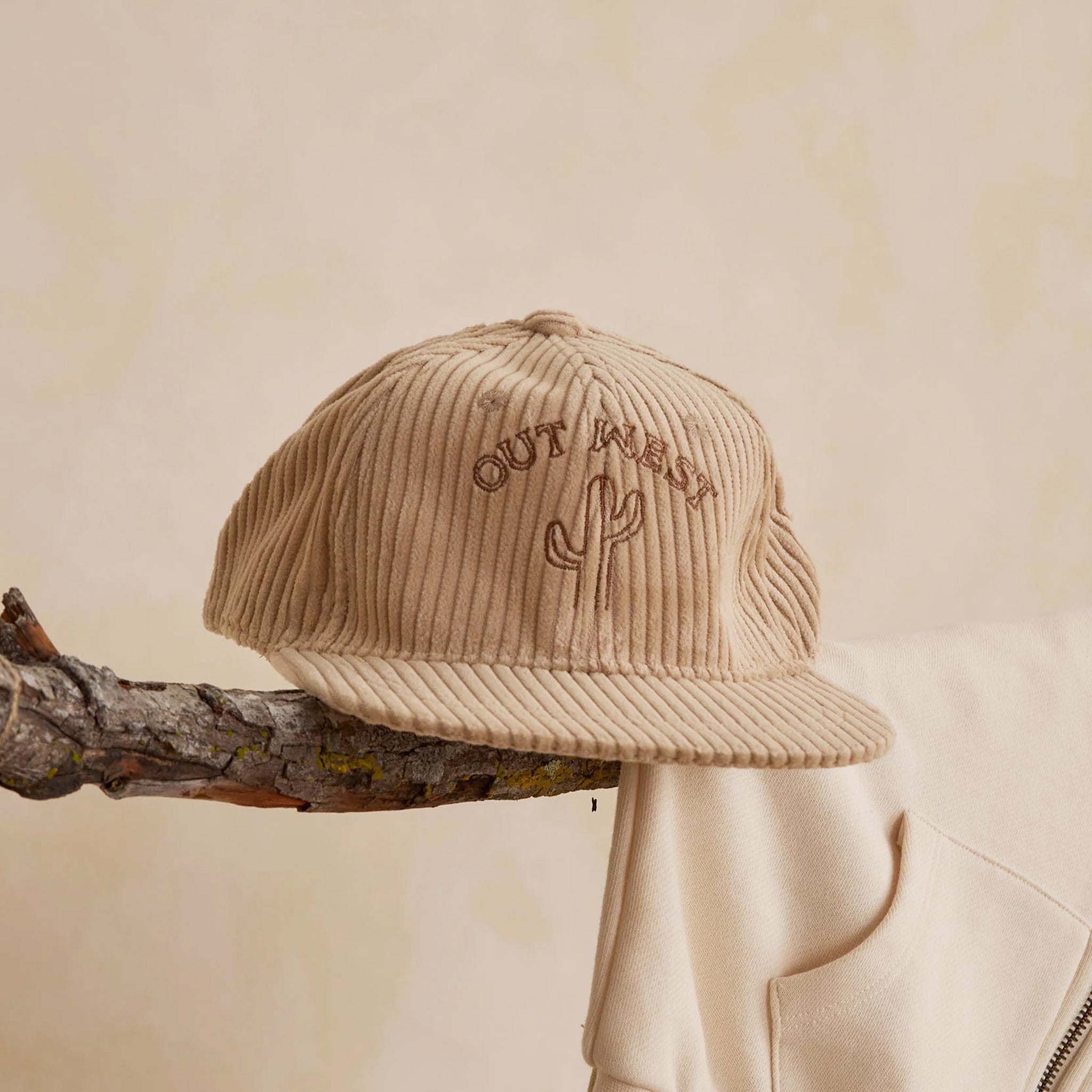 A tan flat brim baseball hat for kids with brown embroidered text on the front that reads, 'Out West' along with a cactus graphic embroidered. 