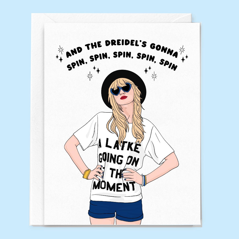 A greeting card sits on a light blue background. The white card with white envelope behind feature an illustration of Taylor Swift standing with her hands on her hips wearing jean shorts and a white t-shirt that reads "A Latke Going on at the Moment".  Above her head the card reads in black font, "And the dreidel's gonna spin, spin, spin, spin, spin". 