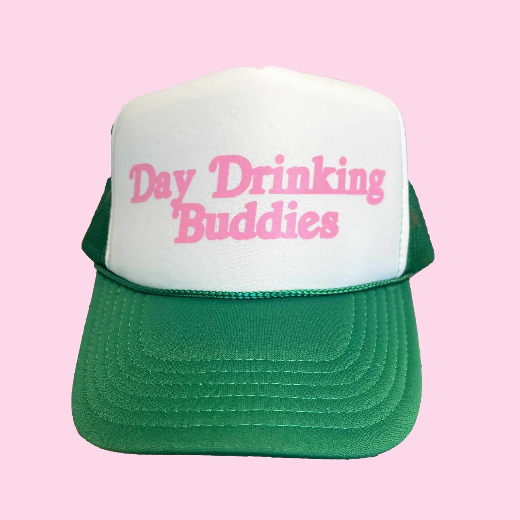 On a pink background is a white and green high brim trucker hat with pink screen printed text that reads, "Day Drinking Buddies". 