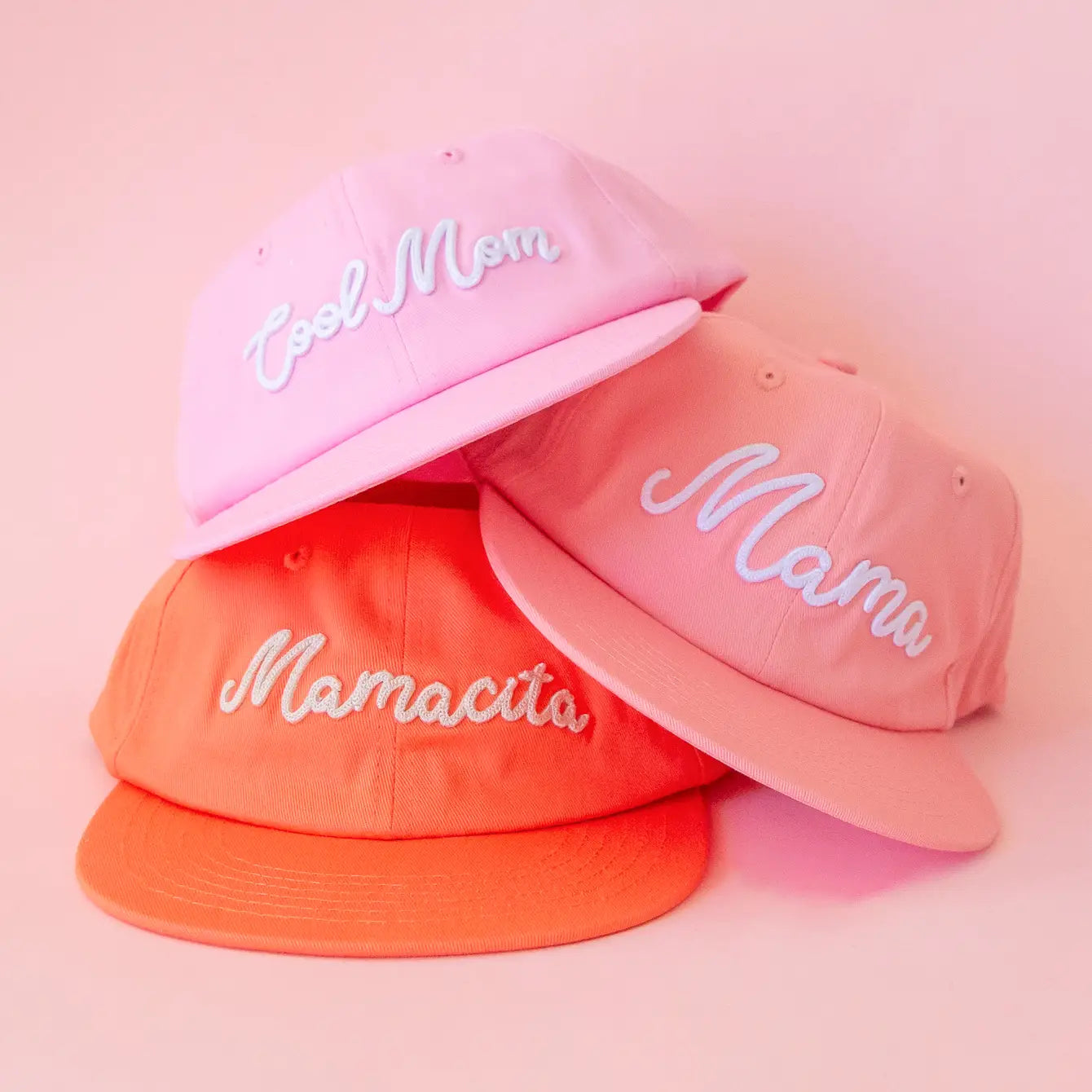 The three pink and orange flat brim hats with different white embroidery on the front reading, 'Cool Mom', 'Mama', and 'Mamacita' on the other. 