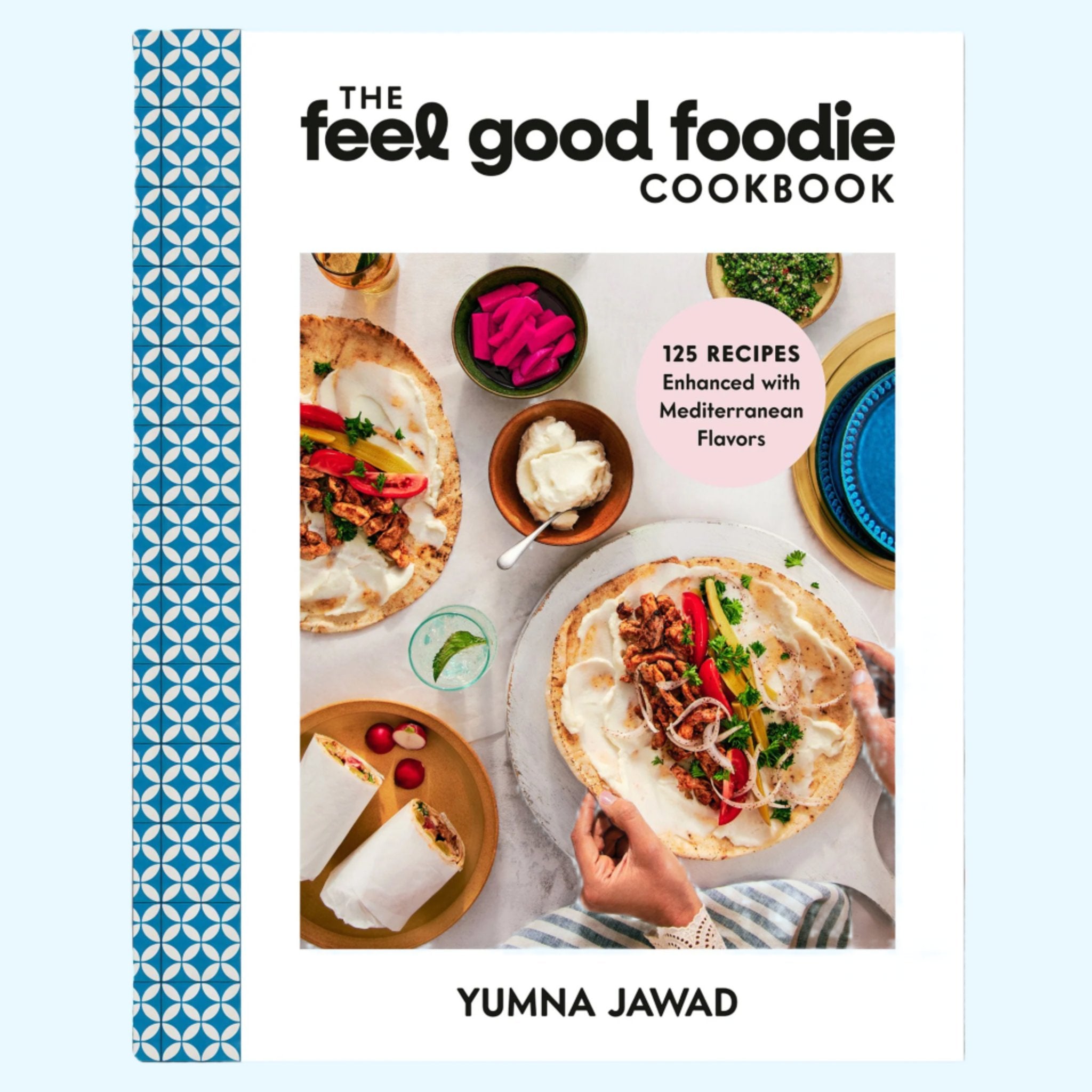 On a light blue background is a white and blue cookbook with a photo of Mediterranean foods on a table and the title that reads, "The feel good foodie Cookbook".