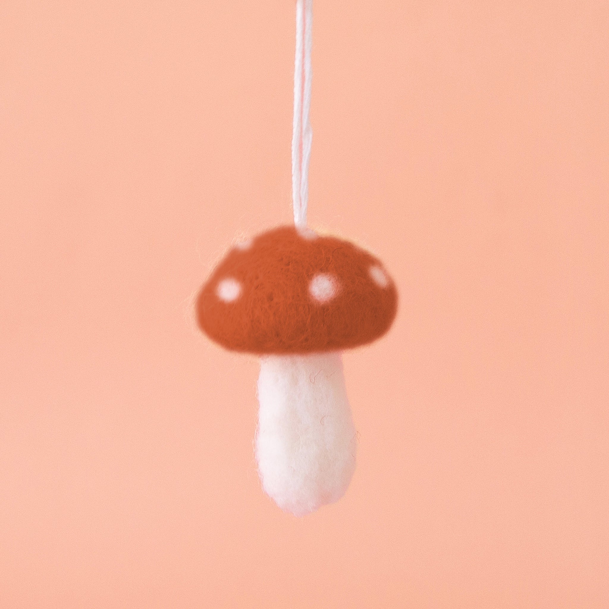 Brown felt mushroom ornament with white spots on the mushroom cap, and a white stem are shown against a peach colored background. 