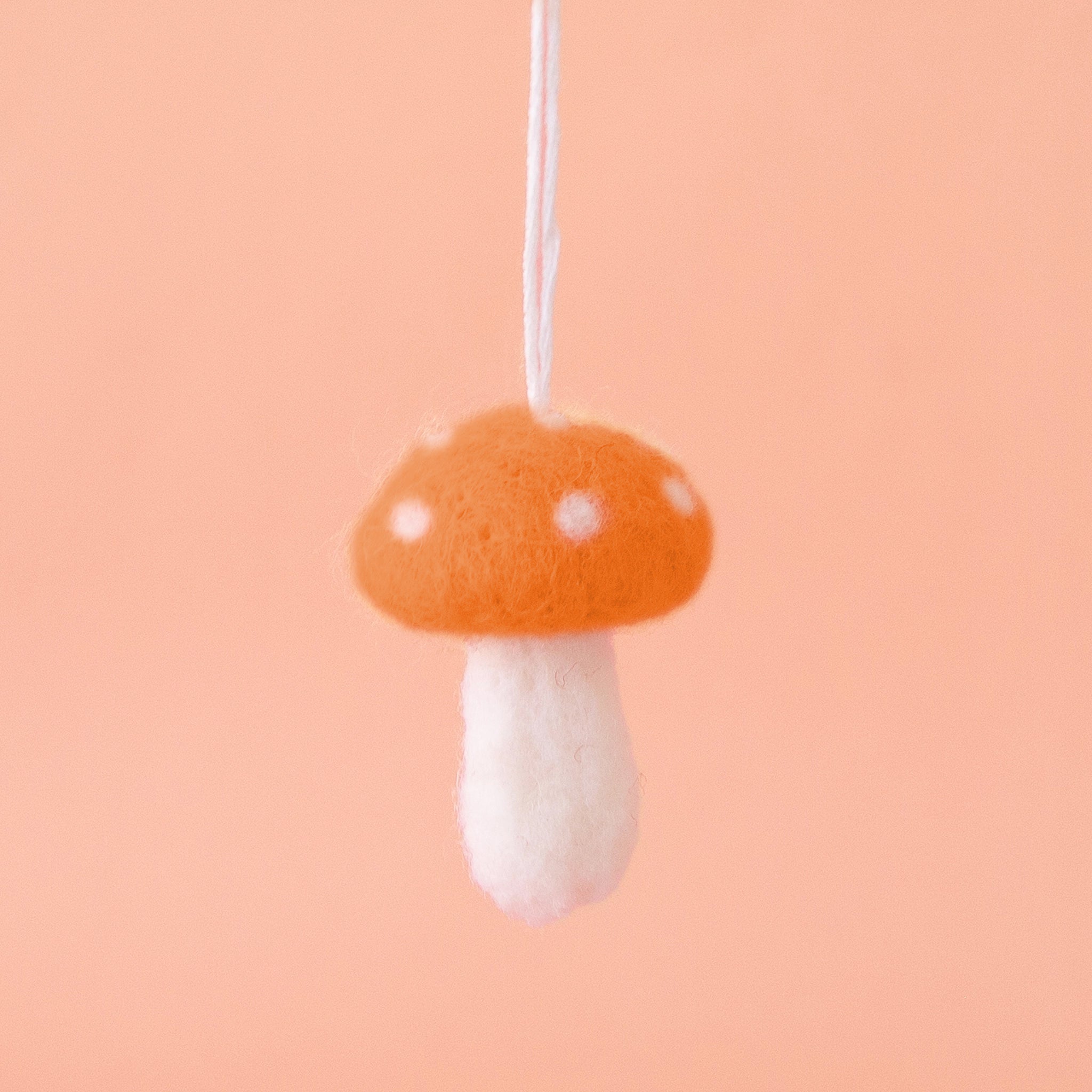 Orange felt mushroom ornament with white spots on the mushroom cap, and a white stem are shown against a peach colored background. 