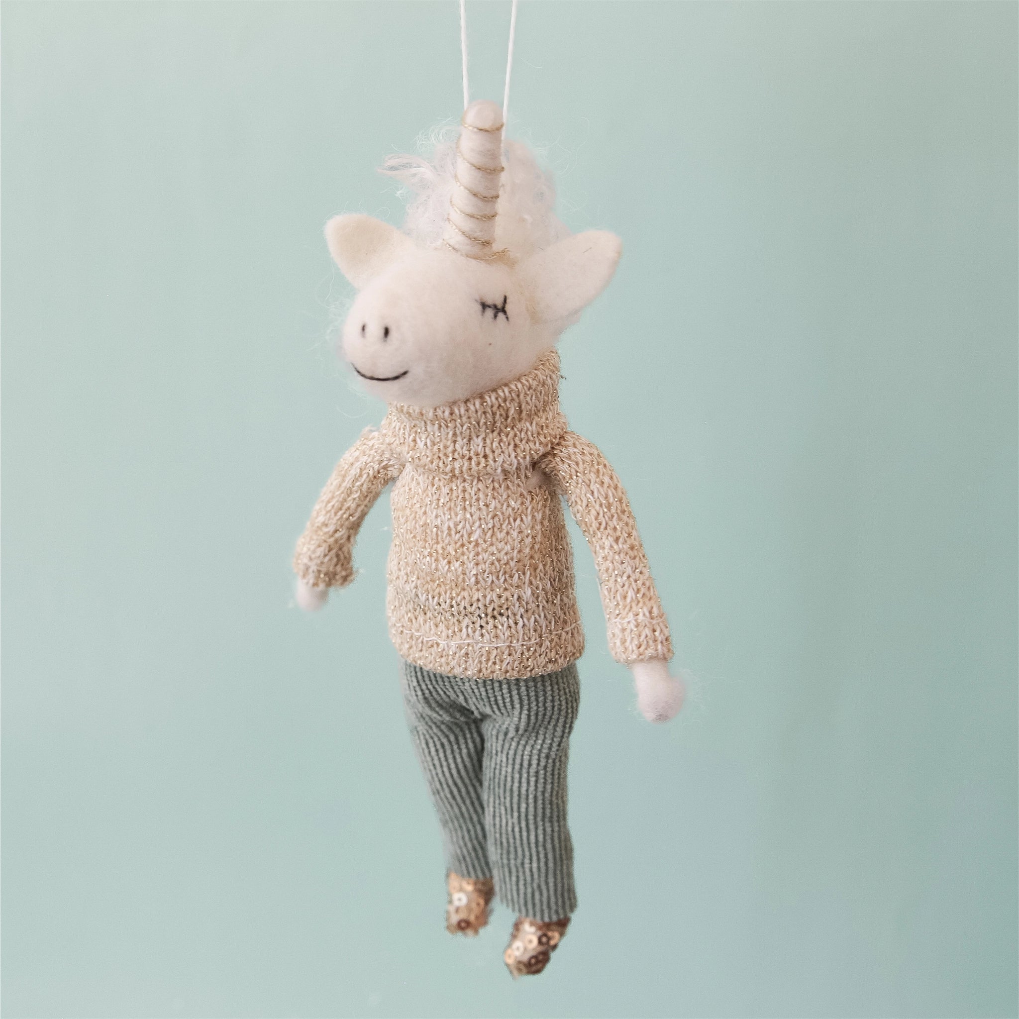 On a aqua blue background is a white felt unicorn ornament with a knit sweater on and corduroy pants and sequin shoes.