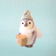 On a blue background is a grey and white felt penguin with a light green beanie and holding a gold gift box in hand. 