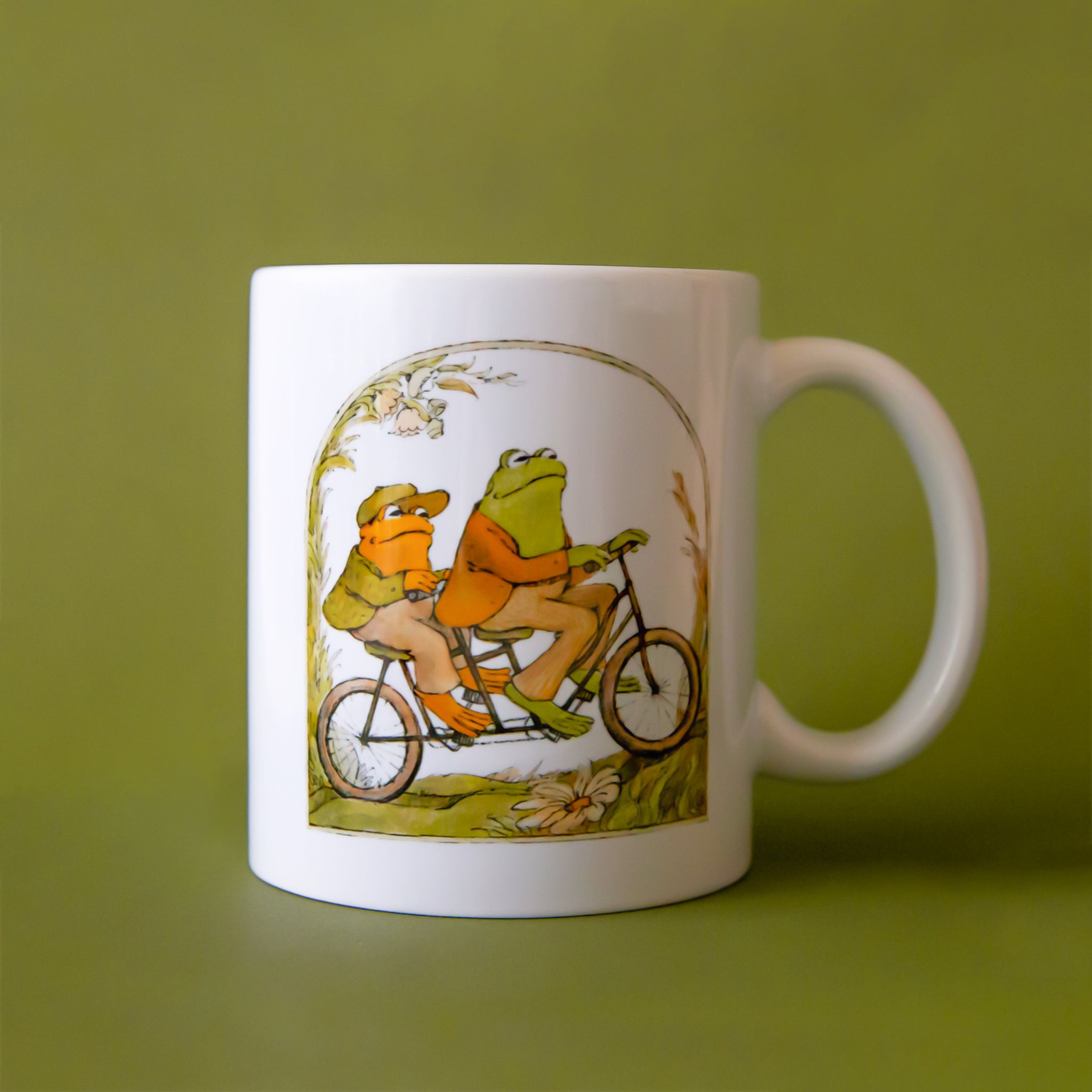 A white mug with an illustration of a frog and toad riding a bike. 