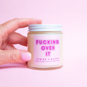 Clear glass jar candle with a white lid off to the side. The label is light pink with font that reads "fucking over it".