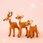 On a pink background is three different sized brown fur reindeer ornaments. There is a baby, a mama and a dad with antlers. Each ornament sold separately. 